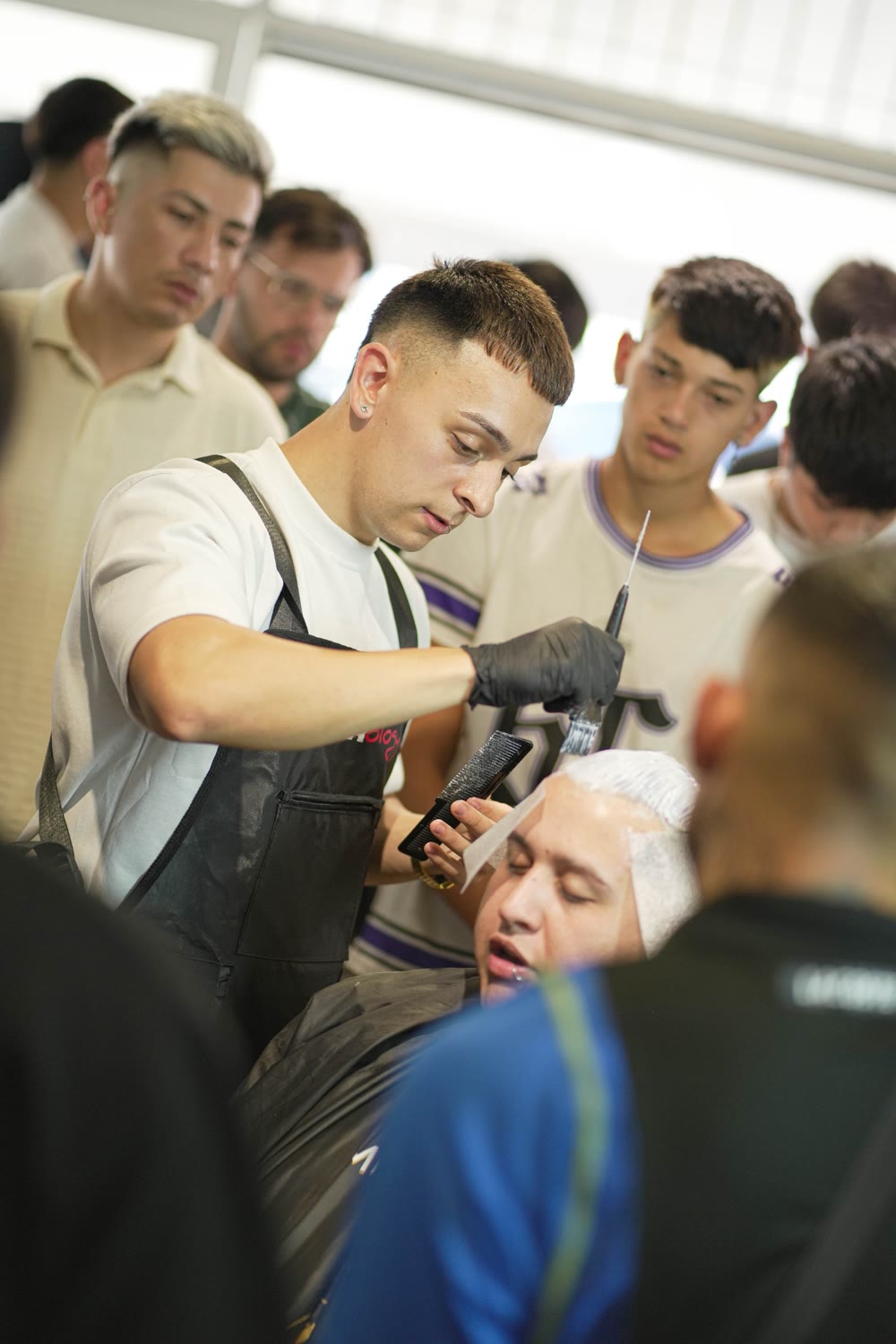Catriel aplicando decolorante en un seminario, rodeado de barberos que miran